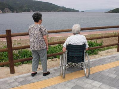 海沿いの遊歩道には点字ブロックが敷かれ、車いすの入居者と付き添いの女性が海を見ている。