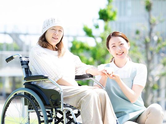車いすの高齢女性にブルーのエプロンを付けた若い女性スタッフが付き添い、屋外で笑顔で過ごしている。