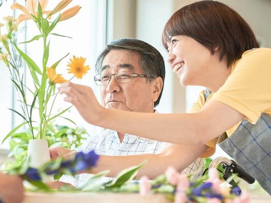 フラワーアレンジメントを楽しんでいる眼鏡をかけた高齢男性は、女性スタッフが手伝っている姿を微笑みながら見ている。黄色や青、ピンクの花が置かれている。