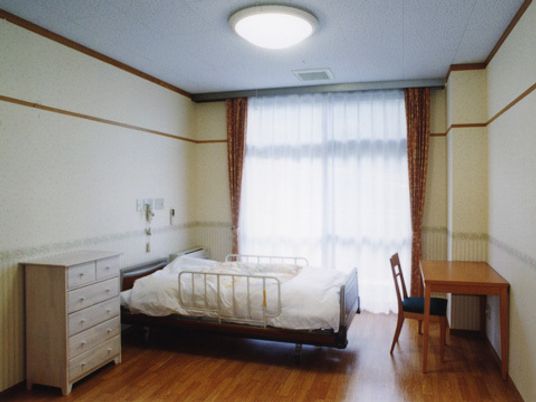寝具付きのベッドのそばには緊急通報装置があり、白いチェストやデスクセットが置かれ、天井には照明があり、大きな窓にはカーテンが付いている。