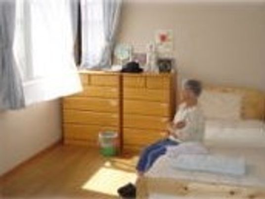 たんすが2つとベッドが置かれた居室。ベッドに座って入居者がくつろいでいる様子。