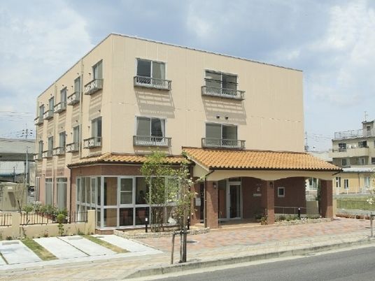 ベージュの外壁の3階建ての建物の前にはレンガでできたアプローチがあり、1階部分には瓦屋根が付いている。