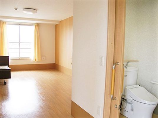 広めの居室内と右手前にあるトイレの様子。左側には日用品入れが置いてある様子