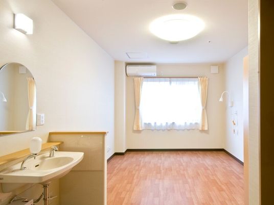レースカーテンとカーテンが付いたおおきな窓があるフローリングの部屋。エアコンや緊急通報装置があり、洗面所も付いている。
