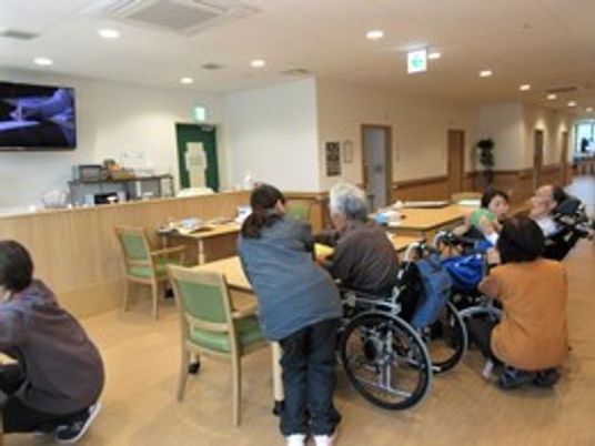 施設内のリビングで車いすの高齢者とスタッフが話をしている様子。日常の一コマ