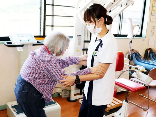 リハビリスタッフが高齢女性の両腕を支え、歩く練習をしている様子。