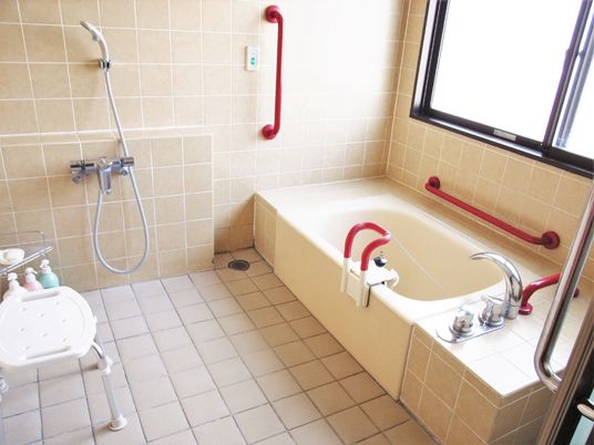 白い浴槽が設置された浴室。シャワーや赤い手すり、シャワーチェアなどもある。