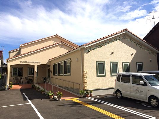 ベージュの外壁にオレンジ系の屋根が付いた明るい雰囲気の建物。駐車場には白線が敷かれ、玄関から手すり付きのスロープが伸びている。