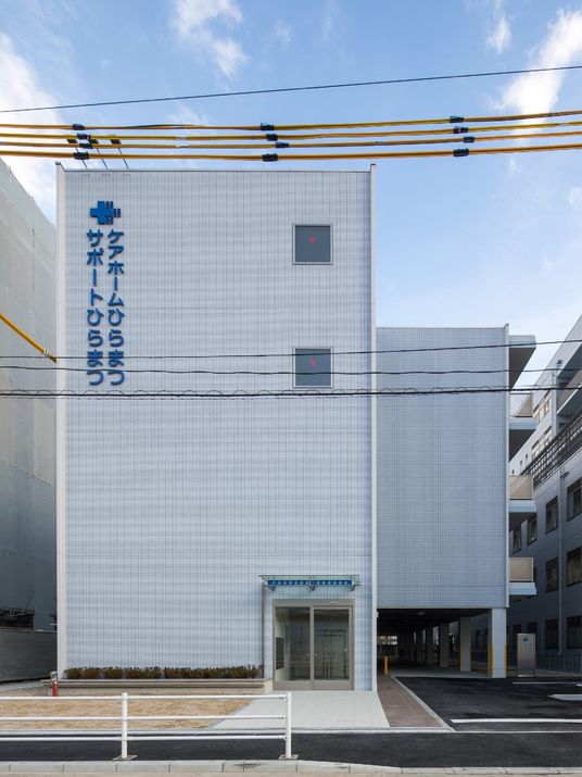 住宅名と併設事業所名が書かれている白い壁が印象的な4階建ての建物。玄関に続く白い道のそばには駐車場がある。