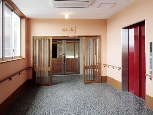 まっすぐな廊下の両サイドに手すりが付き、赤い扉のエレベーターが付き、大きな窓がある。