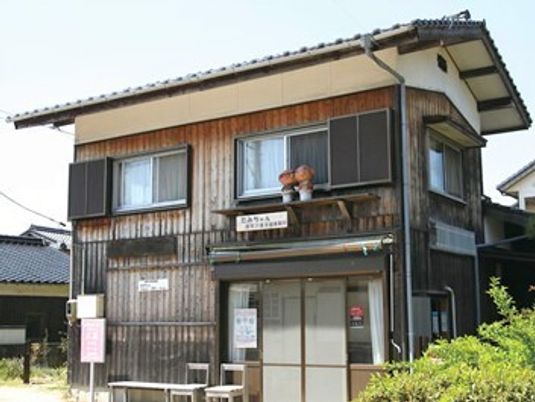 2階建ての木造建築には瓦の屋根が付き、一般の住宅のような外観。玄関先には椅子が2脚とベンチが置かれていて、周囲にも住宅がある。