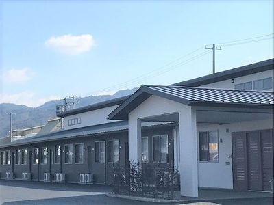 和風の建物は平屋建てで白と黒の落ち着いた色合い。周辺には緑が多く、明るい雰囲気。