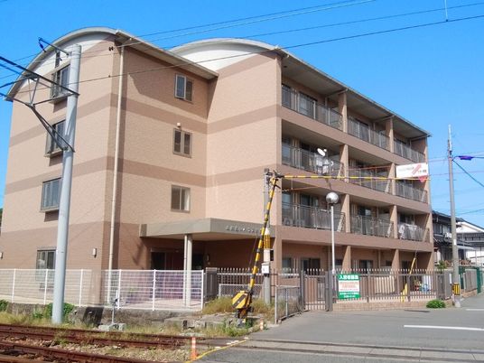 ブラウン系の4階建ての建物の前には踏切があり、角フロアにベランダが付いている。