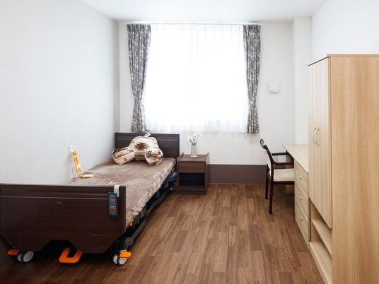 フローリングの部屋には介護ベッドや収納家具、カーテンが付いた窓があり、ベッドサイドには緊急通報装置がある。