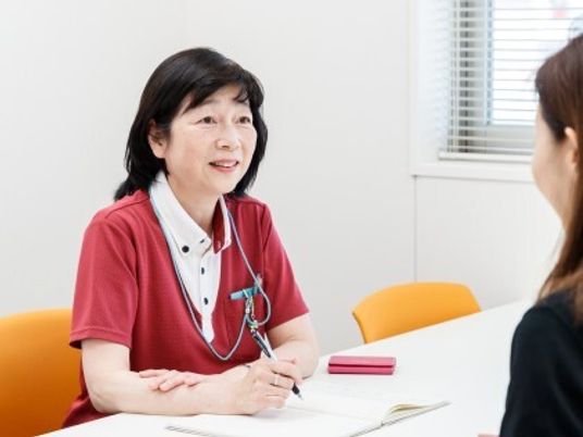 女性スタッフがペンを持ち、メモを取りながら笑顔で髪の長い女性の話を聞いている。