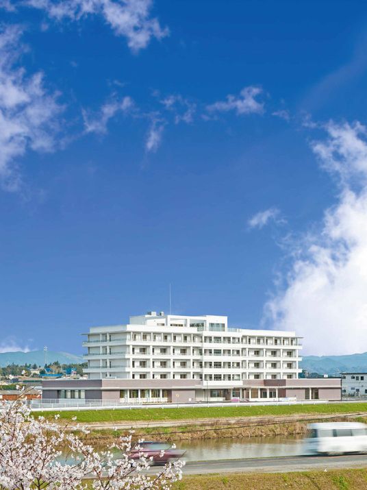 青い空の下に建っている白を基調とした6階建ての建物。川が目の前を流れ、桜の花も咲いている。