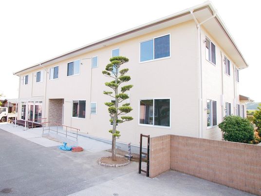 ２階建ての建物はベージュの外壁で玄関先にはスロープが付いていて、手すりも設置されている。周りには木もたくさん植えられている。