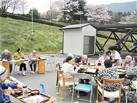 桜並木のそばで食事をとる施設の高齢者の様子。春のお花見の一場面