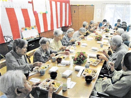 紅白の垂れ幕の前で食事をとっている高齢者の様子。長机に並んで食事をしている写真