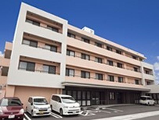 4階建ての建物は横に長く、各フロアにベランダが付き、建物の前には駐車場があり、車が数台止められている。