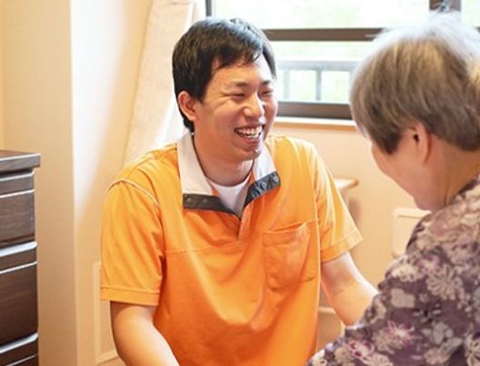 オレンジ色の服を着た男性が、高齢者と目線を合わせて会話をしている