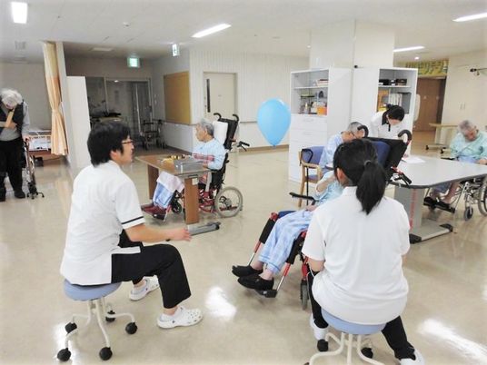 白い服を着た男女が椅子に座っている。目の前には車いすに乗った高齢者が座っている