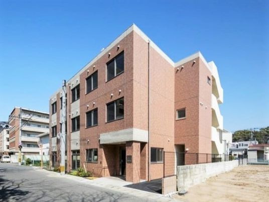 3階建ての建物。ブラウン系で周辺には住宅や緑がある落ち着いた環境。