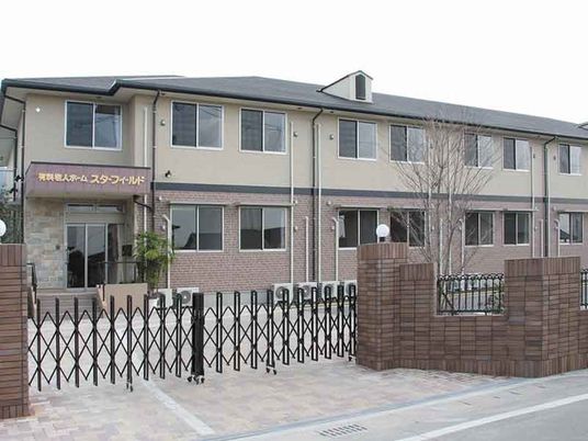 ブラウン系の２階建ての建物。玄関には屋根や階段が付き、門は閉められている。