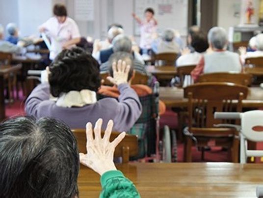 施設内の一室に高齢者が集まってスタッフの指導による体操をしている様子