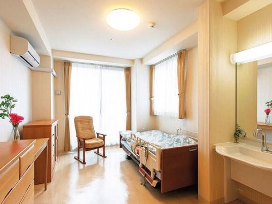 大きな窓にはカーテンが付き、寝具付きのベッドやいす、エアコン、タンス、洗面所などがある部屋。