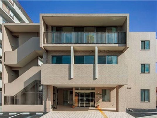 ブラウン系の3階建ての建物。重厚感があり、玄関の前には車寄せがあり、点字ブロックが敷かれ、駐車場もそばにある。