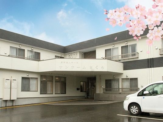 ２階建ての建物は白とグレーのシンプルな色合い。中社長には白い車が止まり、桜の花が咲いている。