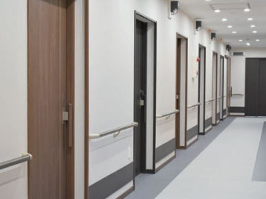 手すりがついて等間隔に部屋の入り口が見えている施設内の廊下の様子