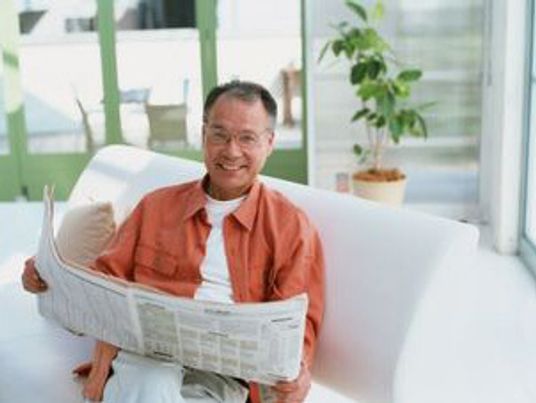 カメラ目線でほほ笑む男性。新聞を読みながらこちらを見ている様子。白いソファに座っている