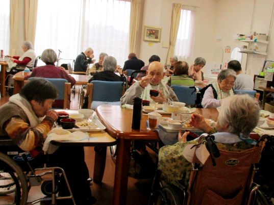 施設内のリビングで食事をとっている高齢者の様子。広めのリビングスペースに多くの人がいる