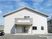 三角屋根が付いた2階建ての建物。白を基調としていて玄関周りだけグレー。玄関の屋根部分にはホーム名が書かれ、駐車場には一台の車が止まっている。