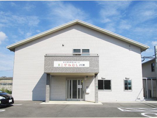 三角屋根が付いた2階建ての建物。白を基調としていて玄関周りだけグレー。玄関の屋根部分にはホーム名が書かれ、駐車場には一台の車が止まっている。