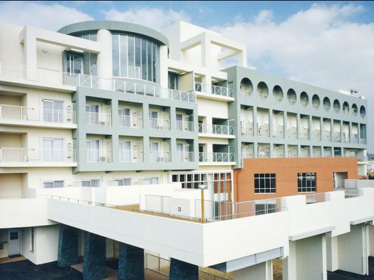 白とグレーの5階建ての建物の角フロアにベランダがあり、日当たりのいい場所に建っている。