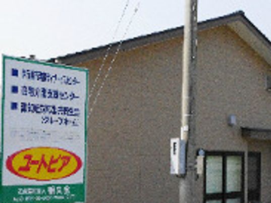 ブラウンの外壁の建物には三角屋根が付いていて、その前にある看板には介護事業所の名前が書かれている。