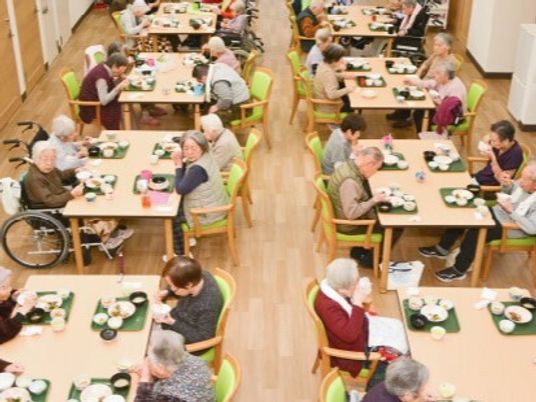 ライトグリーンの椅子がセットされた木製のテーブルが複数並び、入居者が食卓を囲んで食事をしている。