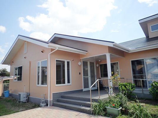 淡いオレンジの外壁が印象的な平屋建ての建物は玄関に手すり付きのスロープと階段が設置され、緑もたくさん植えられている。