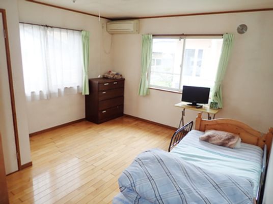 フローリングの部屋には寝具付きの介護ベッドやエアコン、テレビ、タンスなどがあり、大きな2つの窓にはカーテンとレースカーテンが付いている。