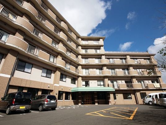 ブラウン系の重厚感のある建物は6階建てで、玄関にはグリーンの屋根が付き、各フロアにベランダが設置され、駐車場には車が止められている。
