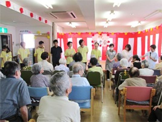 敬老会の紅白の垂れ幕を前にあいさつをするスタッフ。入居者も見守っている。