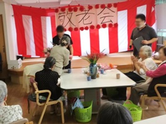 赤い垂れ幕の前で何かを表彰されている人と、拍手をする入居者。