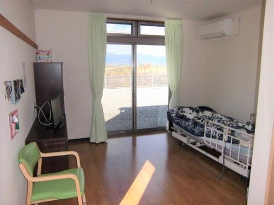 フローリングの居室。ベッドや収納が付き、大きな窓には緑のカーテン、窓の近くにはエアコンがある。
