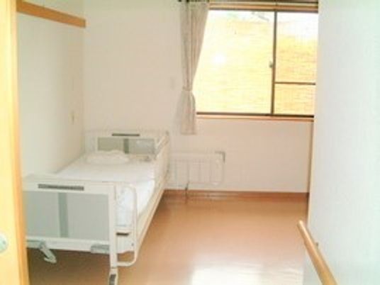 フローリングの居室。ベッドが置かれ、大きな窓にはカーテンがある。