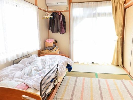 寝具付きのベッドやエアコン、棚などが設置された部屋には大きな窓が二つあり、壁には服もかけられている。