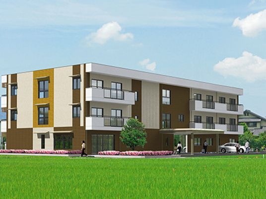 ３階建ての建物はベージュ系の外壁で建物の周りには田畑や花壇があり、民家も見える。