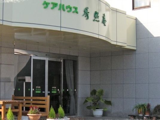 鉄骨造の建物にケアハウス 秀照庵という看板がある様子。手前に木製テーブルと椅子が置いてある様子